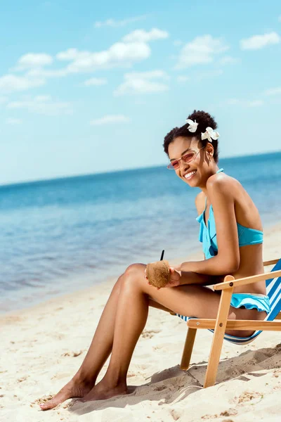 Happy african american woman with cocktail in coconut shell sitting on deck chair on sandy beach — Stock Photo