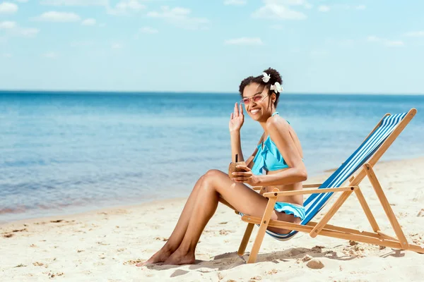 Mujer afroamericana feliz en bikini con cóctel en cáscara de coco saludando a mano mientras se sienta en la silla de cubierta en la playa de arena - foto de stock