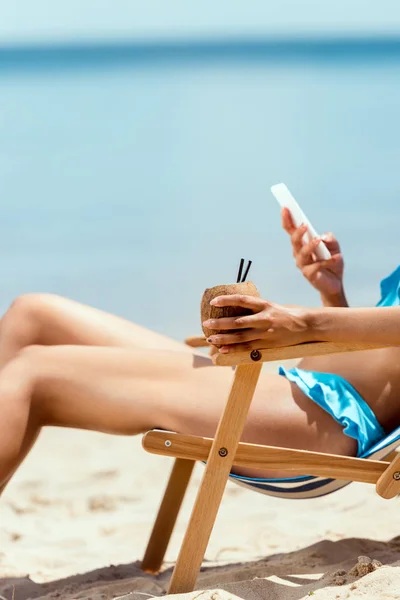 Imagen recortada de la mujer sosteniendo cóctel en cáscara de coco y el uso de teléfono inteligente, mientras que la colocación en la silla de cubierta en la playa de arena - foto de stock