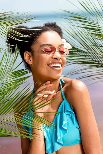 Jovem mulher americana africana sorridente em óculos de sol com flor no cabelo perto de folhas de palma na frente do mar — Fotografia de Stock