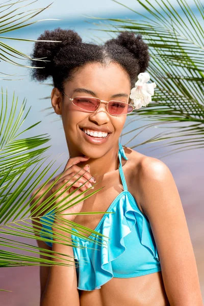 Heureuse femme afro-américaine en lunettes de soleil avec fleur dans les cheveux près des feuilles de palmier en face de la mer — Photo de stock