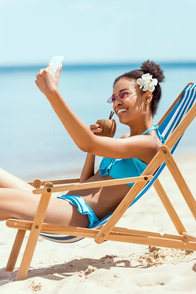 Heureuse femme afro-américaine avec fleur en tête boire un cocktail dans la coque de noix de coco et prendre selfie sur smartphone tout en étant allongé sur une chaise longue sur la plage de sable — Photo de stock