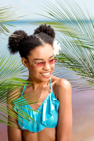 Femme afro-américaine souriante en lunettes de soleil avec fleur dans les cheveux près des feuilles de palmier en face de la mer — Photo de stock