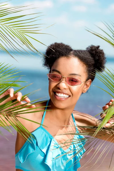 Donna afro-americana sorridente in occhiali da sole vicino alle foglie di palma di fronte al mare — Foto stock