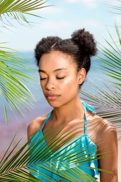 Mulher afro-americana com olhos fechados perto de folhas de palmeira em frente ao mar — Fotografia de Stock
