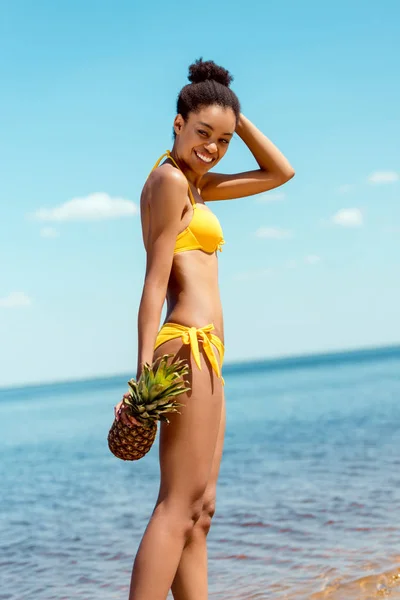 Happy african american woman in bikini holding pineapple on sandy beach — Stock Photo