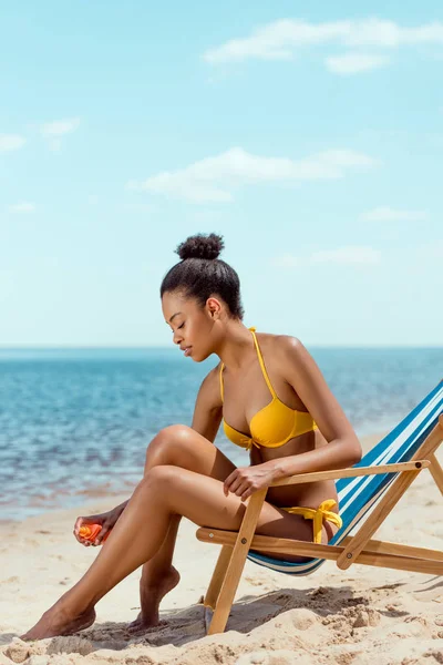 African american woman applying sunscreen lotion on skin while sitting on deck chair on sandy beach — Stock Photo