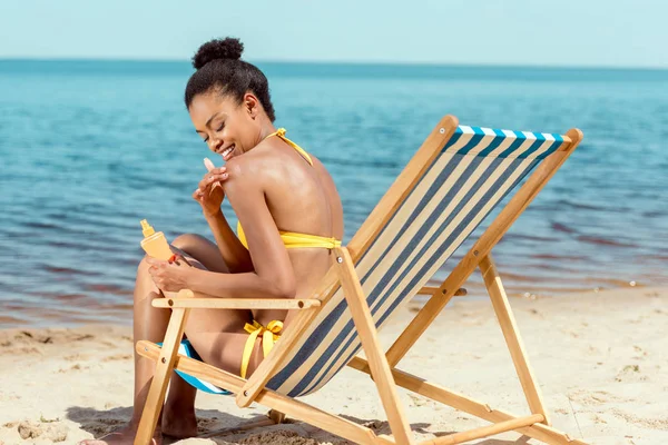 Femme afro-américaine souriante appliquant de la crème solaire sur la peau tout en étant assis sur la chaise longue sur la plage de sable — Photo de stock