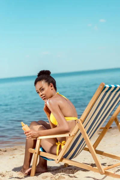 African american woman applying sunscreen lotion on skin while sitting on deck chair on sandy beach — Stock Photo