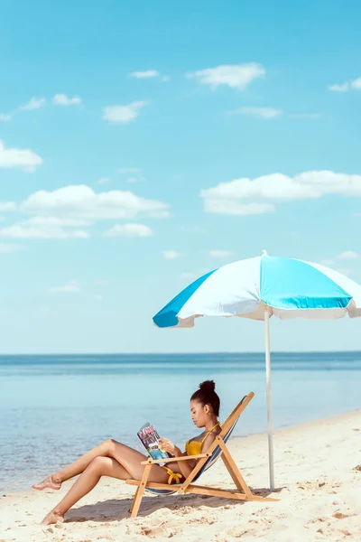 Vista lateral de la joven mujer afroamericana leyendo revista y relajarse en la tumbona bajo el paraguas de la playa en el mar frontal — Stock Photo