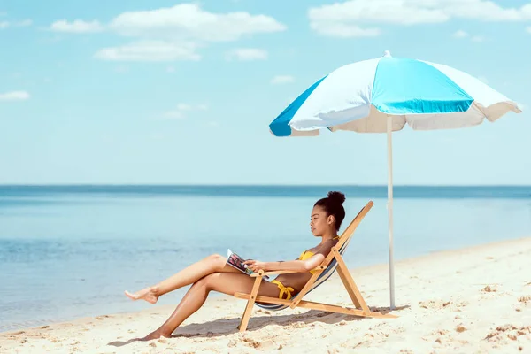 Vista lateral de la mujer afroamericana leyendo revista y relajarse en la tumbona bajo el paraguas de la playa en frente del mar - foto de stock