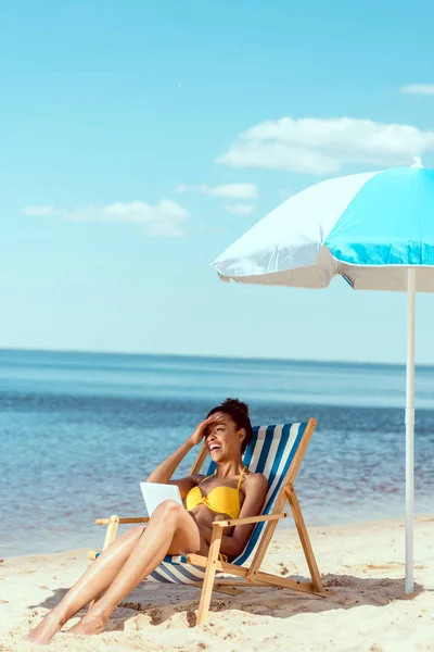 Сміється афроамериканська жінка розслабляється на стільці і використовує цифровий планшет під пляжною парасолькою перед морем — стокове фото