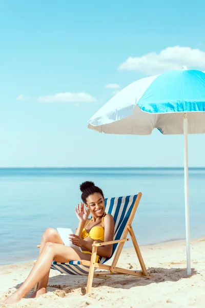 African american woman with digital tablet relaxing on deck chair and waving by hand under beach umbrella in front of sea — Stock Photo