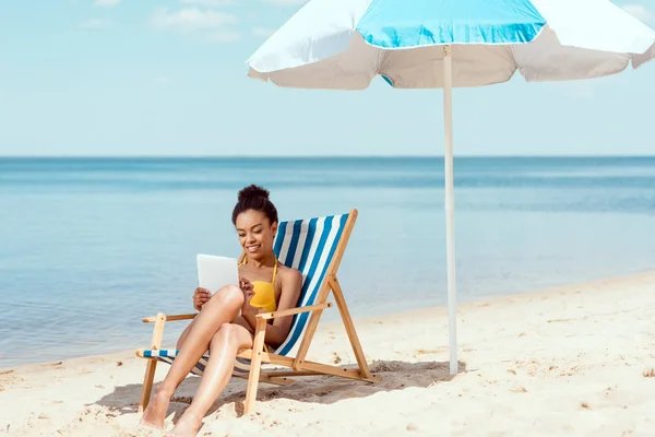 Donna africana americana che si rilassa sulla sedia a sdraio e utilizza tablet digitale sotto l'ombrellone di fronte al mare — Foto stock