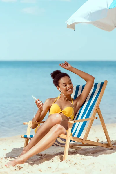 Happy african american woman laying on deck chair and listening music in earphones with smartphone under beach umbrella in front of sea — Stock Photo