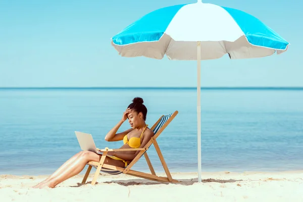 Vista lateral de la sonriente mujer afroamericana freelancer sentado en la silla de cubierta y el uso de la computadora portátil bajo el paraguas de la playa en frente del mar - foto de stock