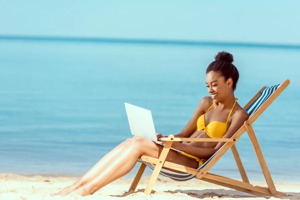 Souriant afro-américaine femme pigiste assis sur la chaise longue et en utilisant un ordinateur portable sur la plage de sable — Photo de stock