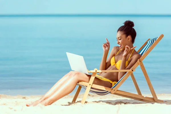 Усміхнена афроамериканка жестикулює пальцями і спілкується ноутбуком, сидячи на стільці на піщаному пляжі — стокове фото