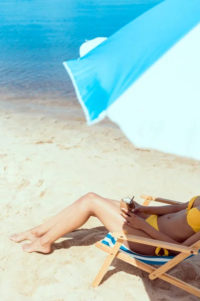 Abgeschnittenes Bild einer Frau im Bikini, die auf einem Liegestuhl liegt und einen Cocktail in Kokosnussschale unter einem Sonnenschirm vor dem Meer hält — Stockfoto