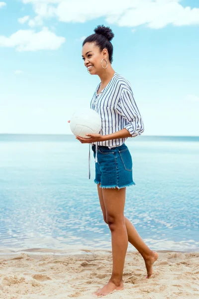 Sonriente mujer afroamericana sosteniendo la pelota de voleibol en la playa de arena - foto de stock