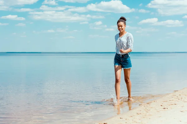 Giovane donna afro-americana sorridente che cammina in acqua di mare vicino alla spiaggia di sabbia — Foto stock