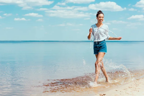 Smiling african american woman running in sea water near sandy beach — Stock Photo