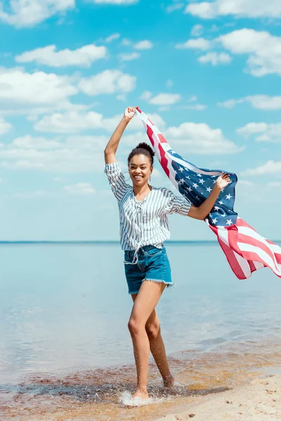 Donna afroamericana sorridente che corre in acqua di mare con bandiera americana, concetto di indipendenza — Foto stock