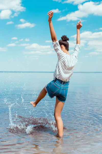 Vista trasera de mujer joven con brazos anchos pateando agua de mar — Stock Photo