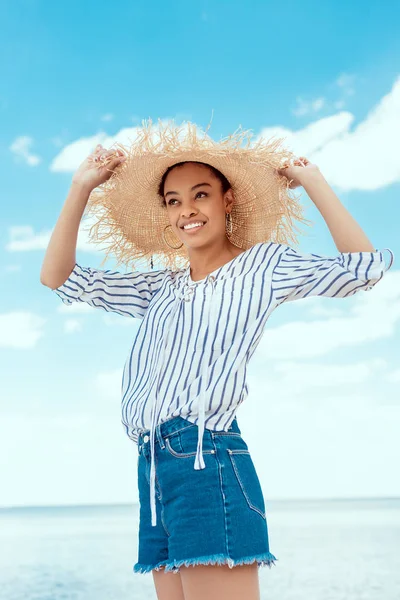 Baixo ângulo de visão da mulher americana africana sorridente em chapéu de palha na frente do mar — Fotografia de Stock