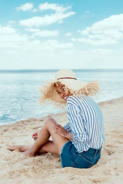 Heureuse femme américaine africaine en chapeau de paille regardant la caméra tout en étant assis sur la plage de sable — Photo de stock