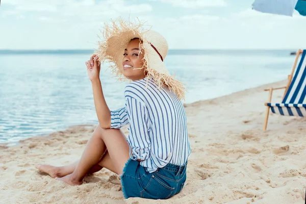 Sonriente mujer afroamericana en sombrero de paja mirando a la cámara mientras está sentado en la playa de arena con tumbona y sombrilla de playa - foto de stock