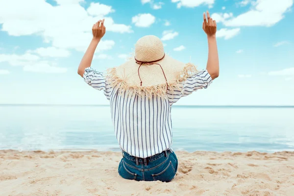 Задний вид женщины в соломенной шляпе с широкими руками, сидящей на песчаном пляже — стоковое фото