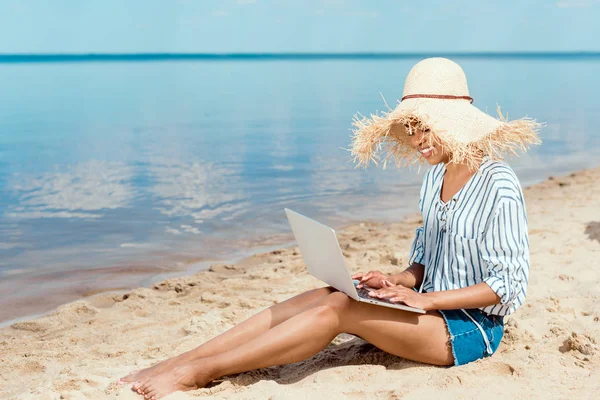 Щаслива афроамериканська фрілансерка в солом'яному капелюсі сидить на піщаному пляжі з ноутбуком — стокове фото