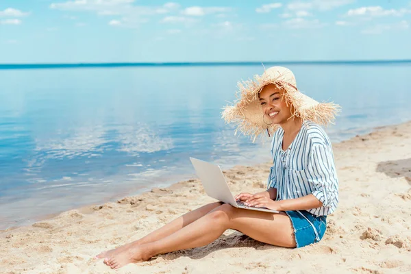 Souriant afro-américaine femme pigiste en chapeau de paille assis sur la plage de sable avec ordinateur portable — Photo de stock