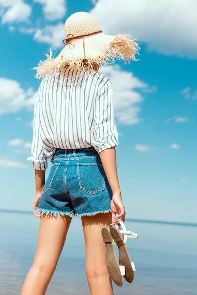 Vista trasera de la mujer en sombrero de paja que sostiene las sandalias y mirando al mar - foto de stock