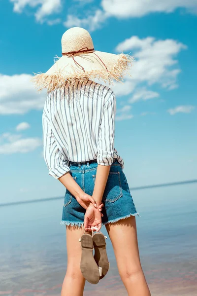 Vista trasera de la mujer en sombrero de paja que sostiene las sandalias y mirando al mar - foto de stock