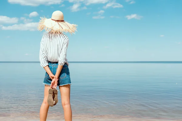 Visão traseira da mulher em chapéu de palha segurando sandálias e olhando para o mar — Fotografia de Stock