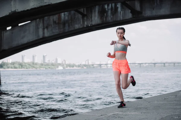Sportive regardant montre intelligente tout en faisant du jogging sur le quai — Photo de stock