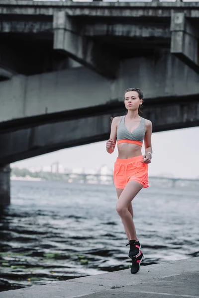 Attractive sportswoman jogging on quay near bridge — Stock Photo