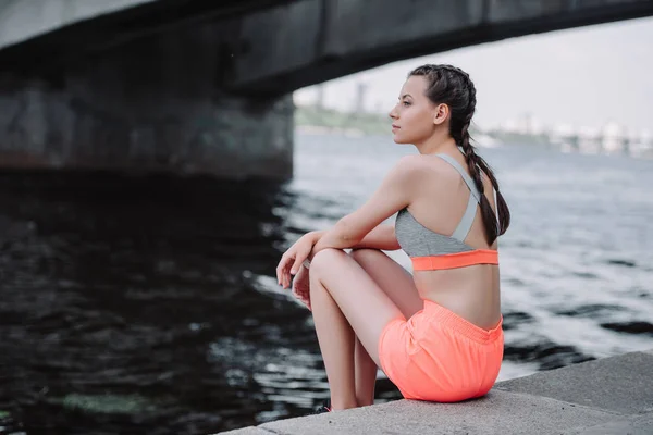 Attrayant jeune femme athlétique en vêtements de sport assis sur le quai — Photo de stock