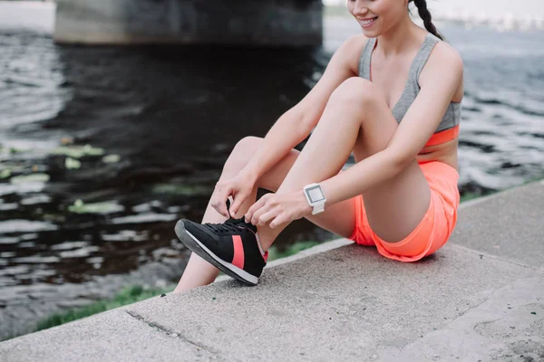 Cropped view of sportswoman tying laces on sneakers and sitting on quay — Stock Photo