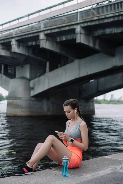 Beautiful sportswoman using smartphone with earphones and smart watch while sitting on quay near bridge — Stock Photo