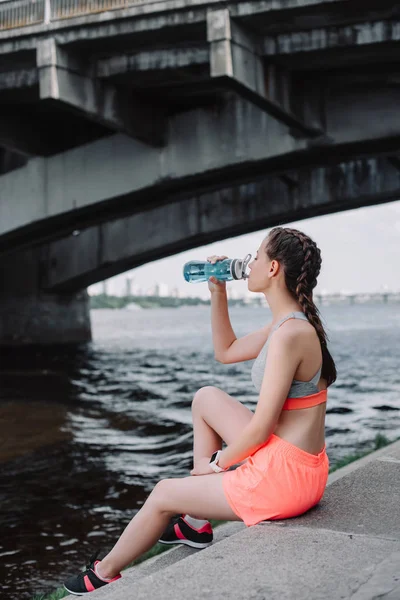 Jovem atlético mulher beber água de esportes garrafa e sentado no cais perto do rio — Fotografia de Stock