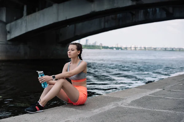 Hermosa deportista con smartwatch y botella de deportes sentado en el muelle cerca del río - foto de stock