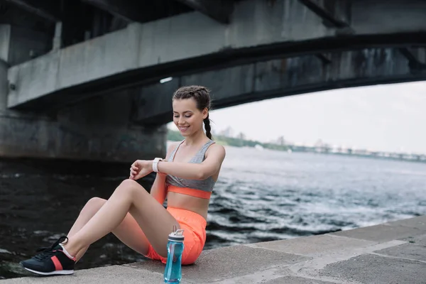 Sportive souriante regardant montre intelligente tout en étant assis sur le quai avec bouteille d'eau — Photo de stock