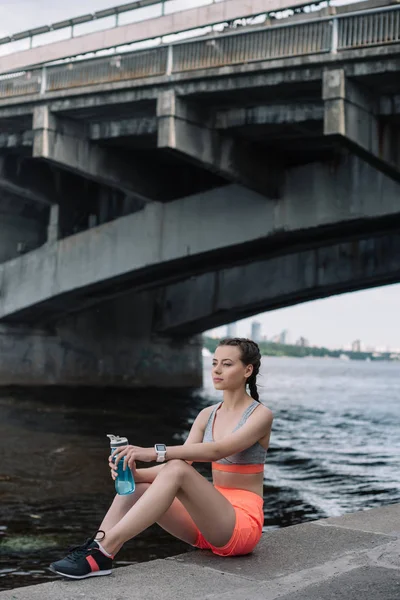 Giovane sportiva con smartwatch e bottiglia sportiva con acqua seduta sul molo vicino al fiume — Foto stock