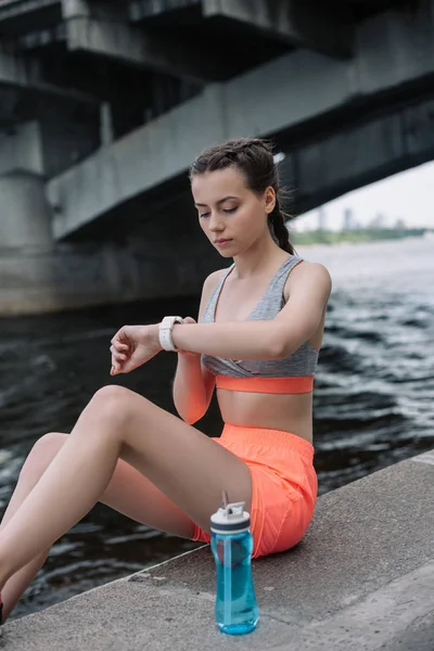Молодая спортсменка смотрит на умные часы, сидя на набережной с бутылкой воды — стоковое фото