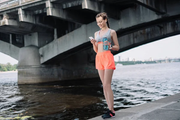 Молода спортсменка тримає смартфон і пляшку з водою — стокове фото
