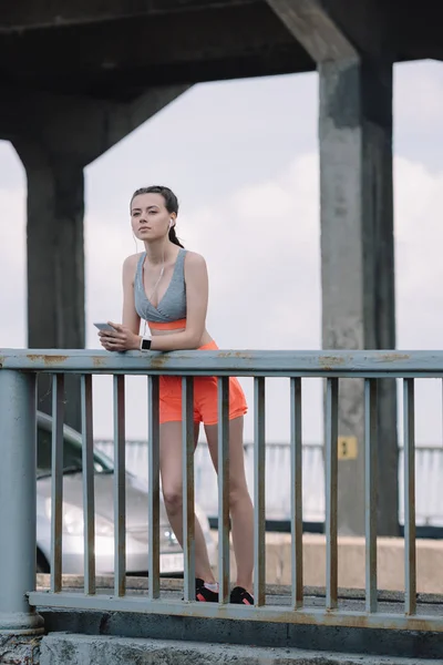 Esportista pensativo com fones de ouvido e smartphone em pé perto de grades na ponte — Fotografia de Stock
