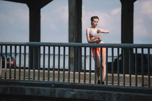 Jovem desportista com smartphone olhando para assistir e de pé na ponte — Fotografia de Stock